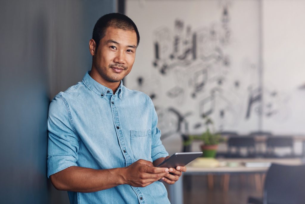 Young Asian man smiling and using a digital tablet while leaning against a gray wall in a modern office