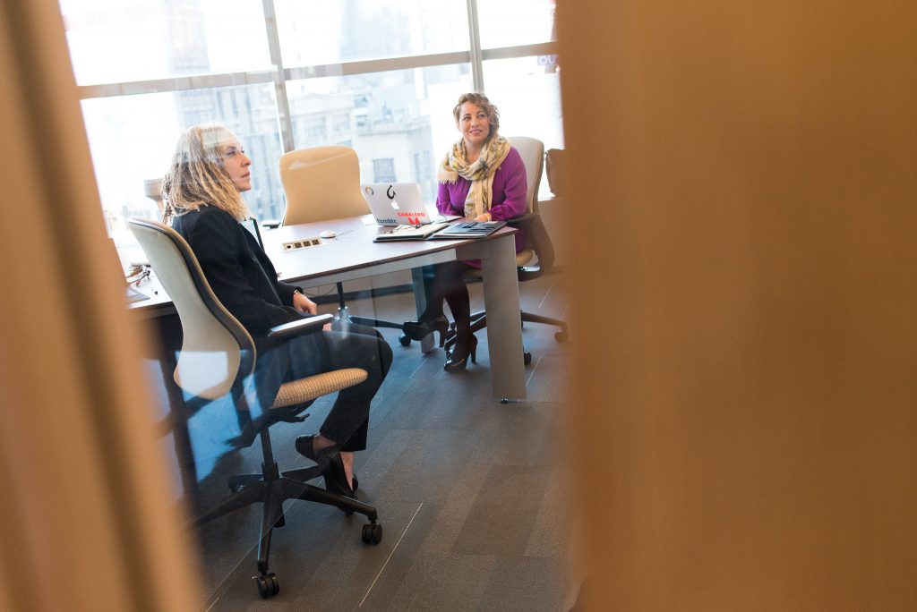 Two woman sitting at an office meeting room table engaged and listening to presenter running training session