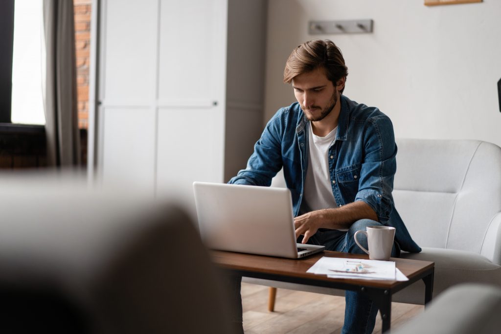 Young man in casual clothing sits on couch at home working at laptop